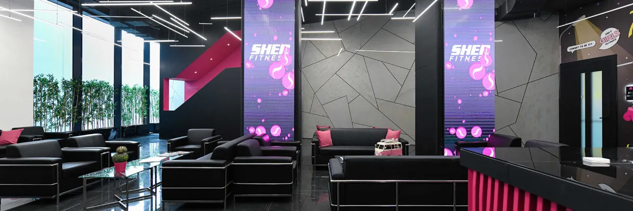 לובי יוקרתי של מועדון הכושר SHE FITNESS עם ריהוט שחור, כריות ורודות, תאורה מודרנית, קירות דקורטיביים ומסכי לד עם הלוגו של המועדון.
